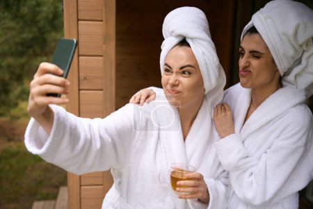 Foto de Mujeres con batas blancas y toallas en las cabezas de pie fuera y sostener la taza, mirando el teléfono inteligente - Imagen libre de derechos