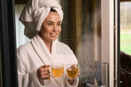 Téléchargez les photos : Dame souriante en manteau blanc et serviette sur la tête debout près de la porte et tenant des boissons - en image libre de droit