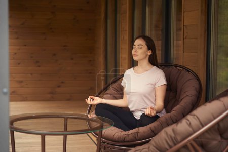 Téléchargez les photos : Femme assise en position lotus sur un fauteuil près de la table, méditer dans le jardin - en image libre de droit