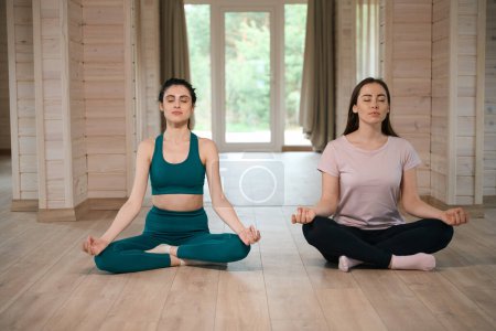 Photo for Young female and trainer in tracksuits meditate next to each other and relaxing - Royalty Free Image