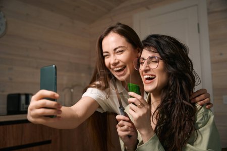 Photo for Smiling female holding smartphone and hugging friend, lady with glasses eating cucumber in the kitchen - Royalty Free Image