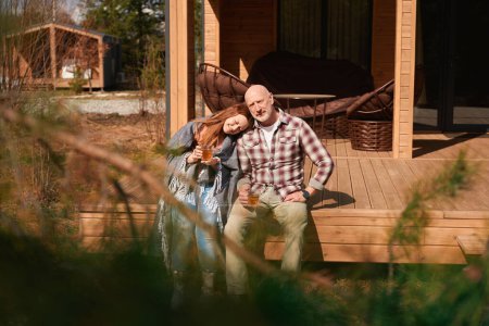 Téléchargez les photos : L'homme et sa femme heureuse assis sur le porche en bois avec des tasses dans les mains - en image libre de droit