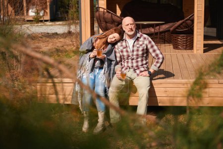 Téléchargez les photos : Femme souriante et son mari sérieux assis sur le porche en bois avec des tasses de thé dans les mains - en image libre de droit