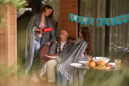 Photo for Smiling male seated in chair on cottage veranda receiving gifts from his family - Royalty Free Image