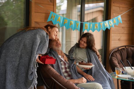 Photo for Birthday person seated in chair on veranda getting presents from cheerful adolescent girl and smiling woman - Royalty Free Image