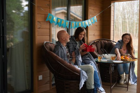 Téléchargez les photos : Adolescente souriante assise sur les genoux de papa déballer cadeau d'anniversaire tandis que sa mère mettre la table à l'extérieur - en image libre de droit