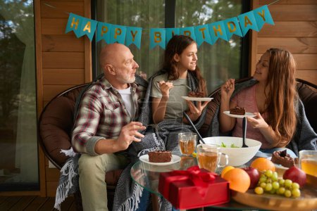 Téléchargez les photos : Joyeux adolescent fille assis à la table de fête sur la terrasse manger gâteau avec ses parents - en image libre de droit