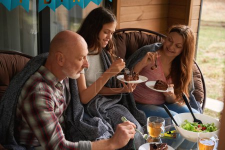 Téléchargez les photos : Famille de trois assis à la table festive sur la véranda chalet manger gâteau au chocolat - en image libre de droit