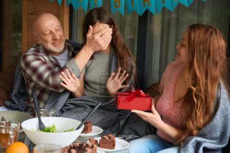 Téléchargez les photos : Père souriant couvrant ses yeux de fille assis à la table festive tandis que sa mère présente un cadeau - en image libre de droit