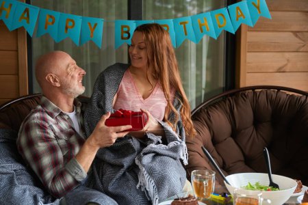 Téléchargez les photos : Gai mature dame assis sur mari tour tout en lui présentant cadeau d'anniversaire sur chalet maison terrasse - en image libre de droit