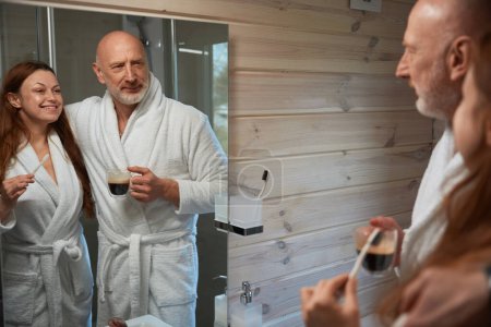 Téléchargez les photos : Homme avec tasse de café et son joyeux compagnon avec brosse à dents à la main en regardant leur reflet dans le miroir - en image libre de droit