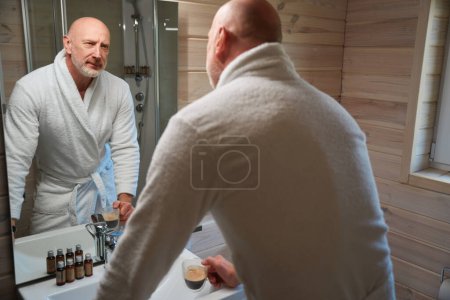 Photo for Serious male with coffee cup leaning on sink while looking at his reflection in mirror - Royalty Free Image