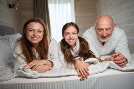 Téléchargez les photos : Adolescente agréable couchée dans un lit confortable avec ses parents heureux - en image libre de droit