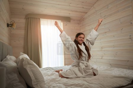 Téléchargez les photos : Joyeux adolescent en peignoir assis sur le lit tout en faisant étirement du bras après le sommeil - en image libre de droit
