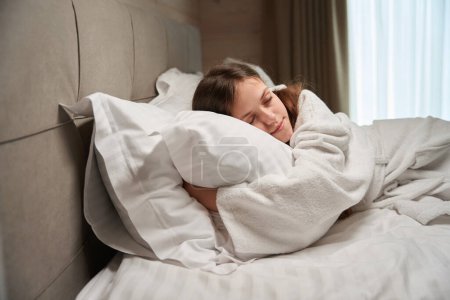 Téléchargez les photos : Jeune femme calme vêtue de peignoir dormant sur un oreiller doux dans un lit confortable - en image libre de droit