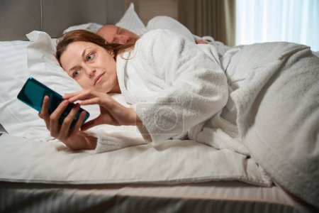 Foto de Mujer madura enfocada leyendo noticias en la pantalla del teléfono inteligente en presencia de su marido en la cama - Imagen libre de derechos