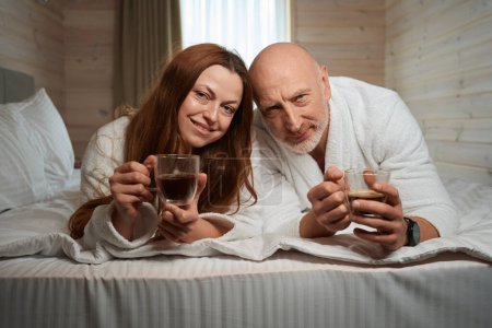 Téléchargez les photos : Conjoints agréables dans des peignoirs couchés dans le lit avec des tasses de café dans les mains - en image libre de droit
