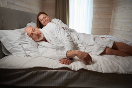 Téléchargez les photos : Dame souriante tapotant son mari sur l'épaule tout en étant couché à côté de lui dans le lit - en image libre de droit