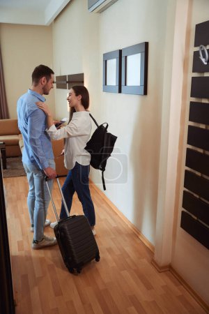 Photo for Smiling female putting her hands on the shoulders of her boyfriend with a suitcase in a hotel room - Royalty Free Image