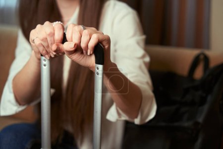 Photo for Lady holding the handle of a suitcase sitting on a sofa in a hotel room. Cropped photo - Royalty Free Image