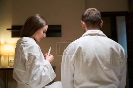 Foto de Mujer joven sentada en la cama junto al hombre en albornoz mirando el dispositivo de prueba de embarazo en sus manos - Imagen libre de derechos