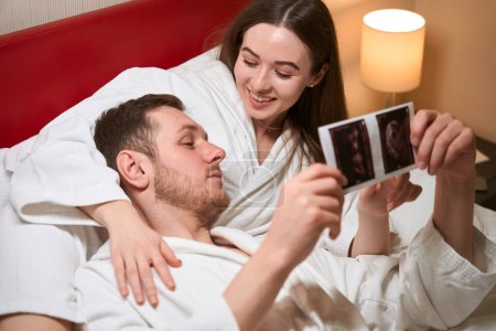Téléchargez les photos : Souriante jeune femme allongée dans son lit dans la chambre d'hôtel tout en pointant le fœtus sur des photos d'échographie à son partenaire - en image libre de droit