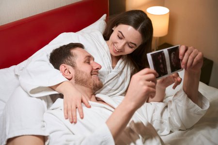 Foto de Feliz joven mujer acostada en la cama en la suite del hotel mientras señala al feto en la ecografía a su complacido compañero - Imagen libre de derechos