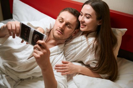 Téléchargez les photos : Femme satisfaite allongée au lit dans la suite de l'hôtel tout en montrant les images échographiques du premier trimestre du bébé au conjoint - en image libre de droit