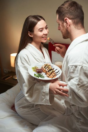 Téléchargez les photos : Homme traitant sa jeune femme heureuse avec un délicieux petit déjeuner dans leur lit - en image libre de droit