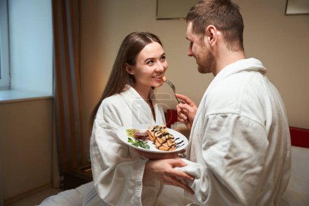 Photo for Loving male treating joyful young female companion with delicious pancakes in bed - Royalty Free Image