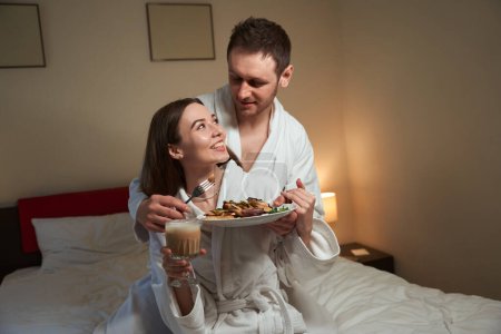 Foto de Cuidar joven alimentación sonriente cónyuge femenino con sabrosos buñuelos en la cama - Imagen libre de derechos