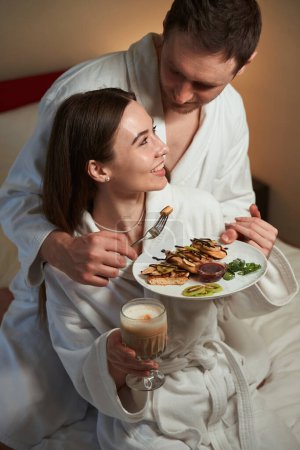Foto de Romántico macho tratando a su feliz compañera femenina con tortitas en suite de hotel - Imagen libre de derechos