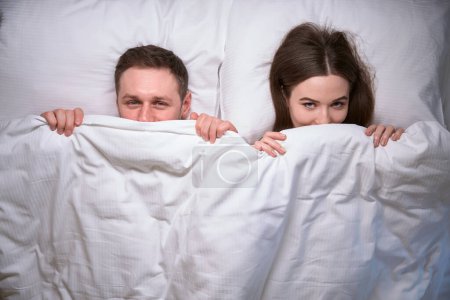 Téléchargez les photos : Un jeune homme et une jeune femme souriants couvrant leur visage avec une couverture alors qu'ils étaient couchés dans leur lit - en image libre de droit