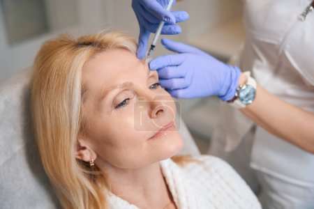 Foto de Primer plano de manos cosmetólogas realizando inyección rejuvenecedora para alisar arrugas en cara de dama madura en salón de belleza - Imagen libre de derechos
