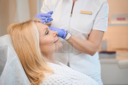 Photo for Close up of cosmetologist hands performing rejuvenating injection for smoothing wrinkles on mature lady face in beauty salon - Royalty Free Image