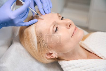 Foto de Primer plano de manos cosmetólogas haciendo inyección rejuvenecedora para suavizar las arrugas en la cara de dama madura en el salón de belleza - Imagen libre de derechos