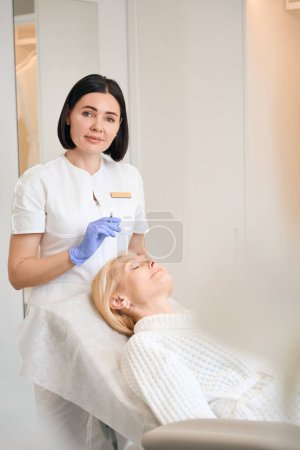 Téléchargez les photos : Cosmétologue heureux tenant la seringue au-dessus du visage de femme mature dans le salon de beauté - en image libre de droit