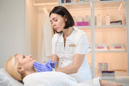 Téléchargez les photos : Cosmetologue faire une injection sur le menton de femme mature dans le bureau d'esthéticienne - en image libre de droit