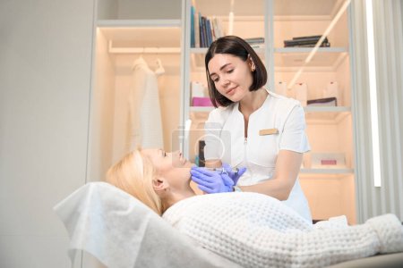 Téléchargez les photos : Cosmétologue souriant faisant une injection sur le menton de femme mature dans un bureau d'esthéticienne - en image libre de droit