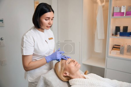 Téléchargez les photos : Cosmetologue faire injection sur femme mature intersourcil dans le salon de beauté - en image libre de droit