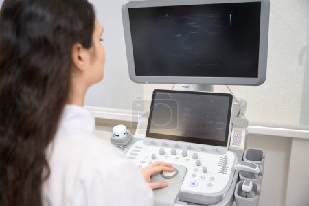 Photo for Young woman cardiologist sitting near the ultrasound scanner in cabinet in the hospital - Royalty Free Image