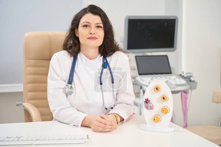 Téléchargez les photos : Jeune femme médecin en manteau blanc assise à table près de la maquette à la clinique - en image libre de droit