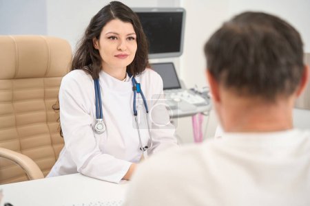 Foto de Mujer joven doctora en bata blanca sentada en la mesa y mirando a la paciente en el hospital - Imagen libre de derechos