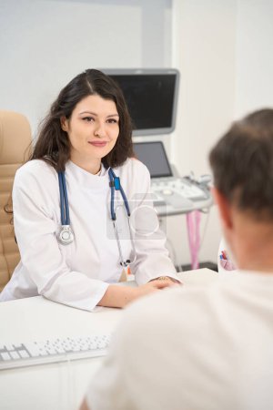 Photo for Young female cardiologist in white coat sitting at table and looking at man in the clinic - Royalty Free Image