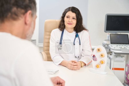 Foto de Mujer doctora sentada en la mesa cerca de la maqueta y mirando al hombre en la sala de examen de ultrasonido en el hospital - Imagen libre de derechos
