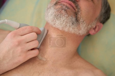 Téléchargez les photos : Médecin tenant scanner et effectuant un examen échographique de la glande thyroïde de l'homme en clinique - en image libre de droit