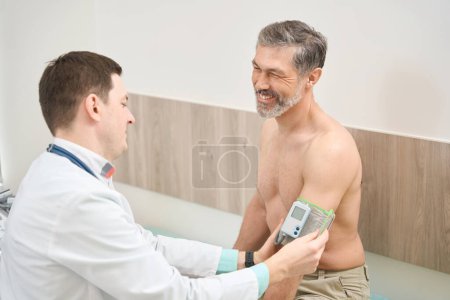 Photo for Physician in white coat sitting near smiling man and measuring pressure in the hospital - Royalty Free Image