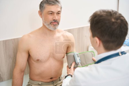 Physician in white coat sitting near male and measuring pressure in the clinic