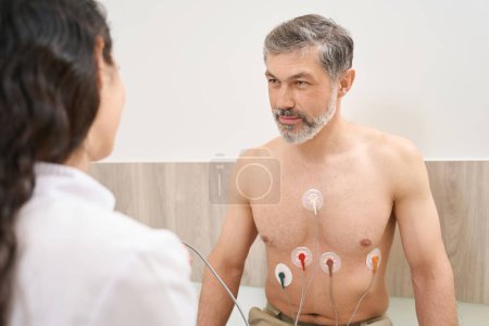 Male sitting near nurse and undergoes an electrocardiography of heart in the hospital