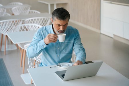 Téléchargez les photos : Homme assis sur une chaise à table et tenant une tasse, tapant sur un ordinateur portable dans la salle à manger - en image libre de droit